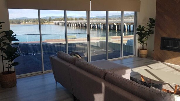 A modern living room with a large window overlooking a body of water and a bridge. There is a sofa, a small table, and two potted plants. - The Lakehouse Windang (Windang)