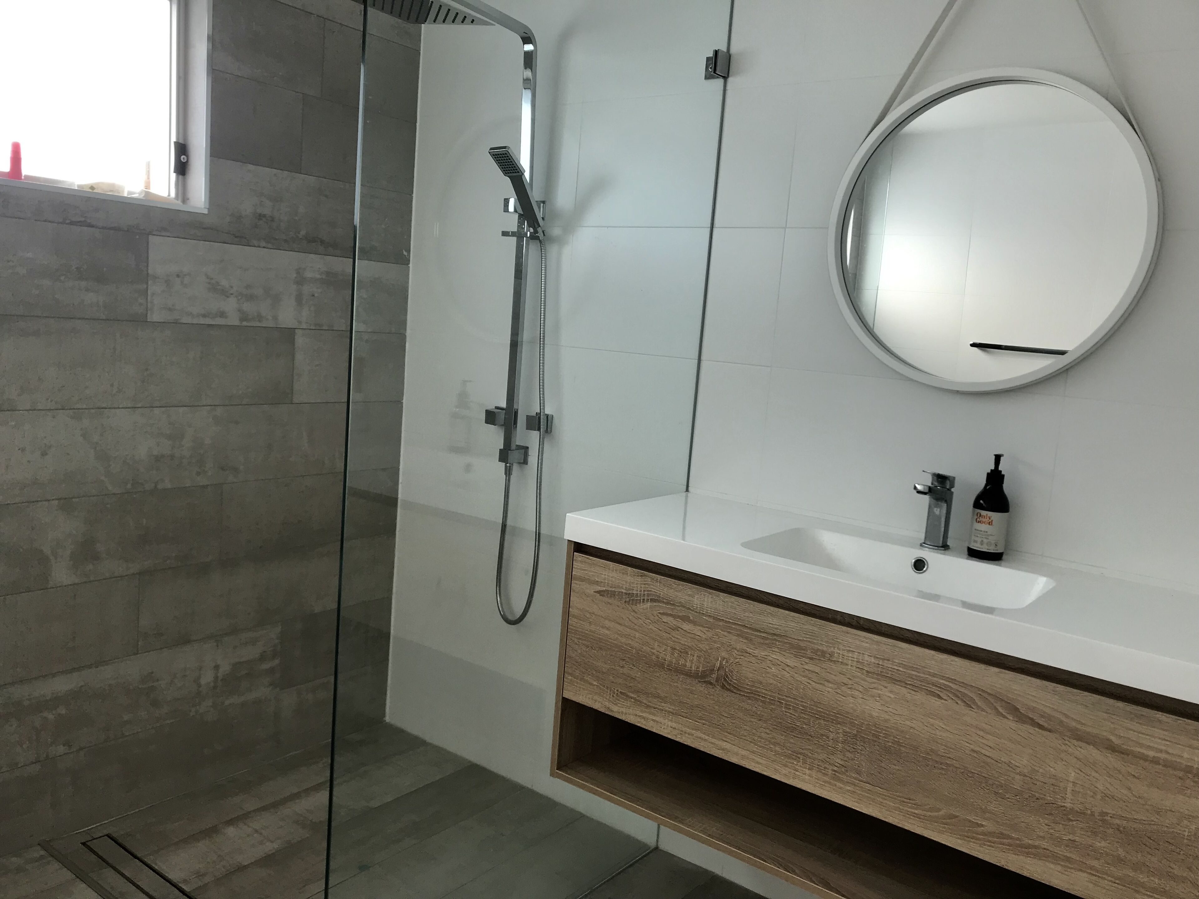 Une salle de bain moderne avec une douche à l’italienne en verre, un lavabo blanc avec un meuble-vasque en bois et un miroir rond au mur.