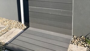 A modern exterior with a grey panel wall, a metal door with two round handles, and a small step leading to a gravel area. - The Lakehouse Windang (Windang)