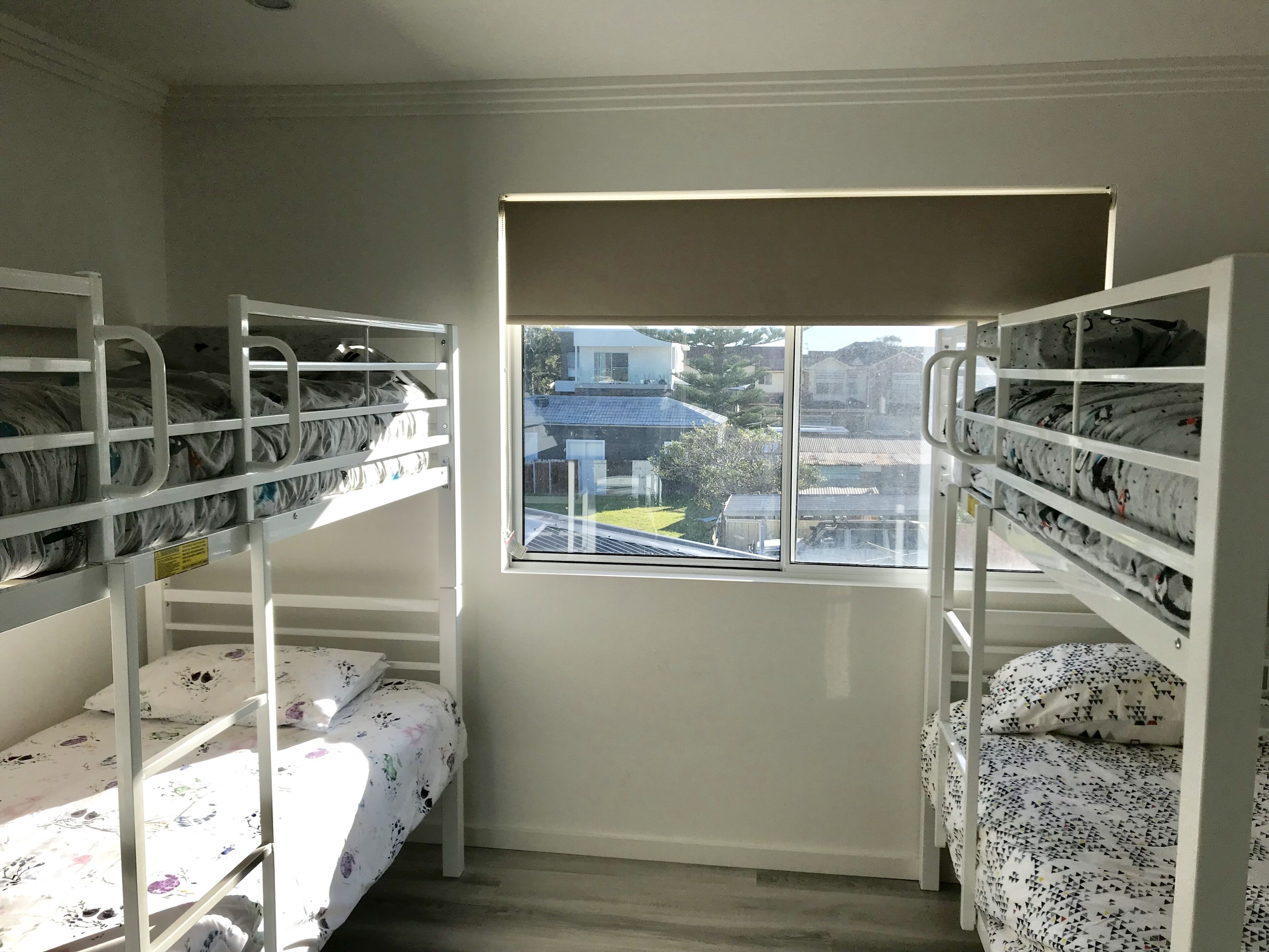 A dormitory room with bunk beds, a window with a view of houses, and a door.