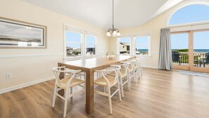 A spacious dining area with a long wooden table and white chairs, large windows with a view, and a balcony with outdoor seating. - Glistening Sands | Oceanfront | Private Pool, Hot Tub | Nags Head (Nags Head)