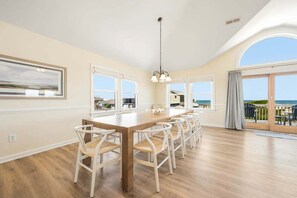 A spacious dining area with a long wooden table and white chairs, large windows with a view, and a balcony with outdoor seating.