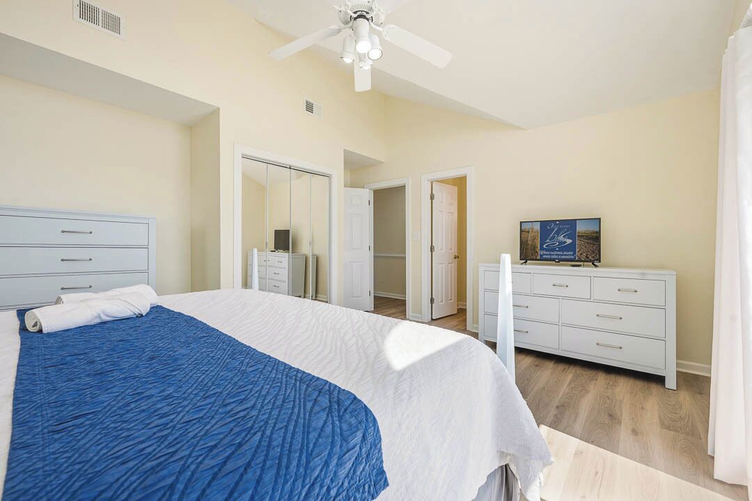 A bedroom with a bed, a dresser, a ceiling fan, and a door leading to another room.