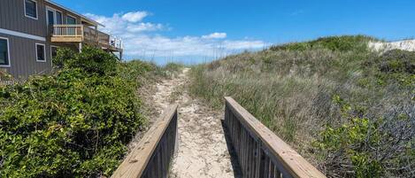 A wooden boardwalk leading to a building with a balcony, surrounded by sand dunes and vegetation.