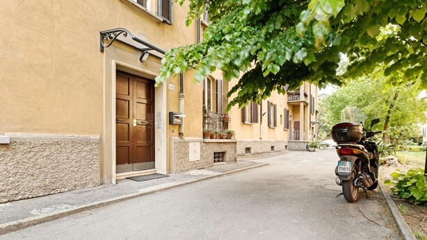 A narrow street with a parked motorcycle, a building with a brown door, and a small porch with potted plants.