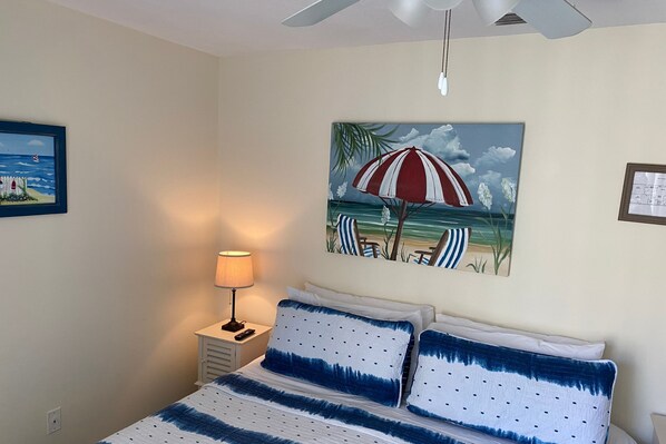 Um quarto com uma cama, uma mesa de cabeceira, um abajur e uma pintura de uma praia.