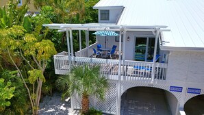 A covered outdoor deck with blue chairs and a table, surrounded by lush greenery and a palm tree.