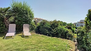Zwei Liegestühle auf einer grünen Wiese mit Blick auf ein Gebäude und üppiges Grün im Hintergrund.
