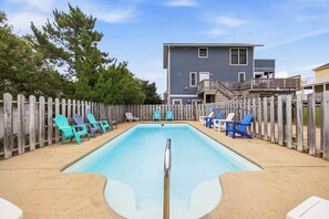 A rectangular swimming pool with a wooden deck surrounding it, a wooden fence, and a two-story building in the background.