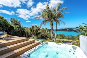A hot tub with a wooden deck and a view of a coastal landscape with palm trees and a clear blue sky.