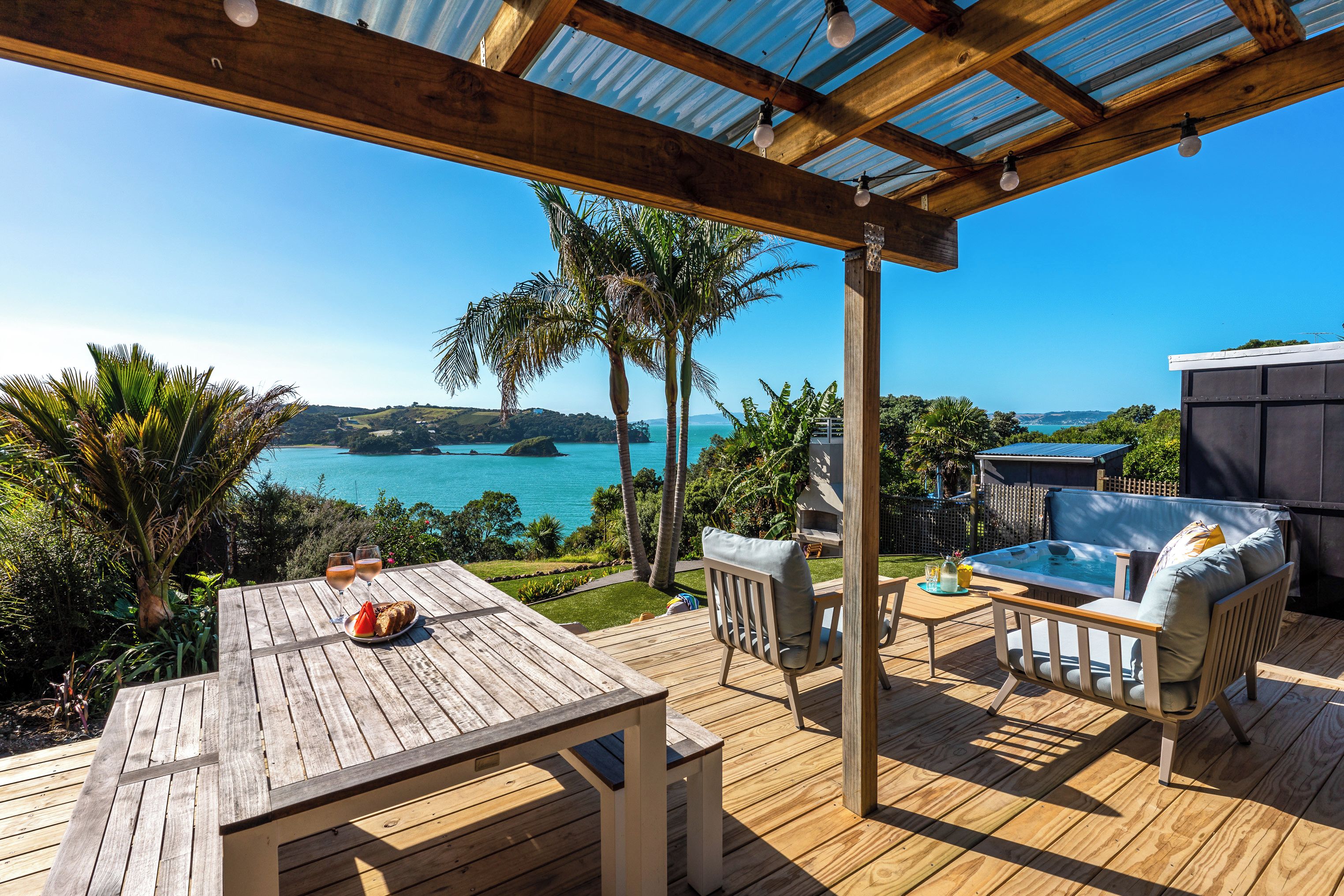 A wooden deck with a table, chairs, and a hot tub overlooking a turquoise sea.