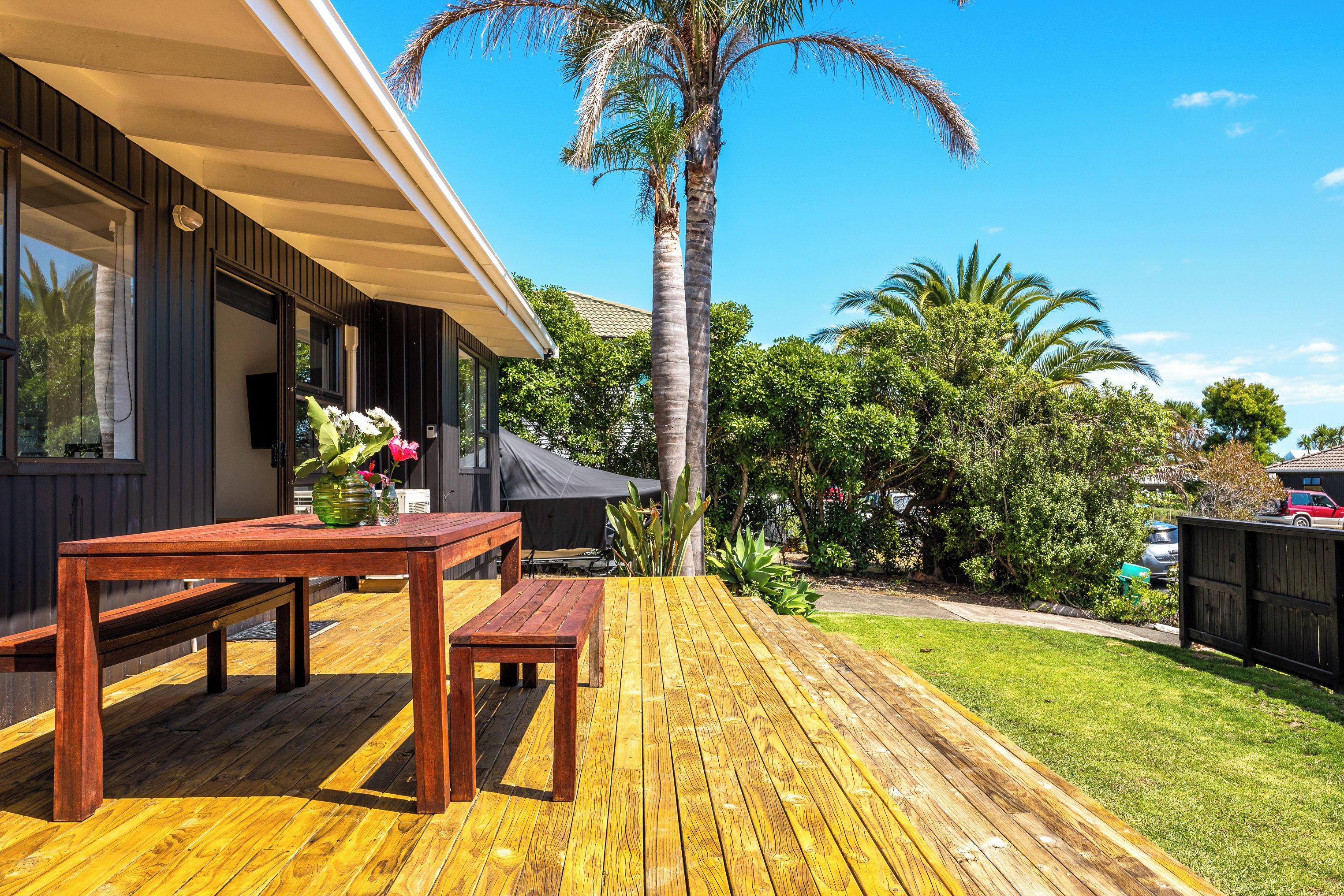 A wooden deck with a bench and table, surrounded by lush greenery and a tall palm tree.