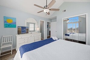 Une chambre à coucher comprenant un lit, un ventilateur de plafond, une commode, une chaise, un tableau et une porte donnant sur une autre pièce.