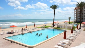 A pool area with lounge chairs and umbrellas, a beach with umbrellas, and a building with balconies. - Beautifully decorated and completely remodeled with custom woodwork throughout... close proximity to geo thermal pools. NO PETS (New Smyrna Beach)