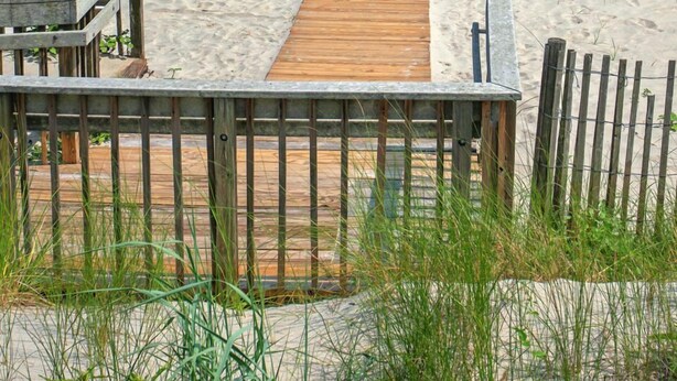 En strand med en träpromenad som leder ner till vattnet, en sandstrand och parasoller i bakgrunden.