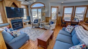 A cozy living room with a fireplace, comfortable seating, and large windows. - Fair Winds | 376 ft from the beach | Private Pool, Hot Tub | Nags Head (Nags Head)