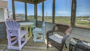 A covered outdoor area with a table and chairs, a wicker chair, and a view of the landscape. - Fair Winds | 376 ft from the beach | Private Pool, Hot Tub | Nags Head (Nags Head)