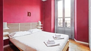 Ein Schlafzimmer mit Bett, Nachttisch, Fenster mit Balkonblick und einer Tür.