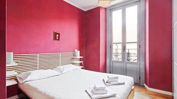 Ein Schlafzimmer mit Bett, Nachttisch, Fenster mit Balkonblick und einer Tür.