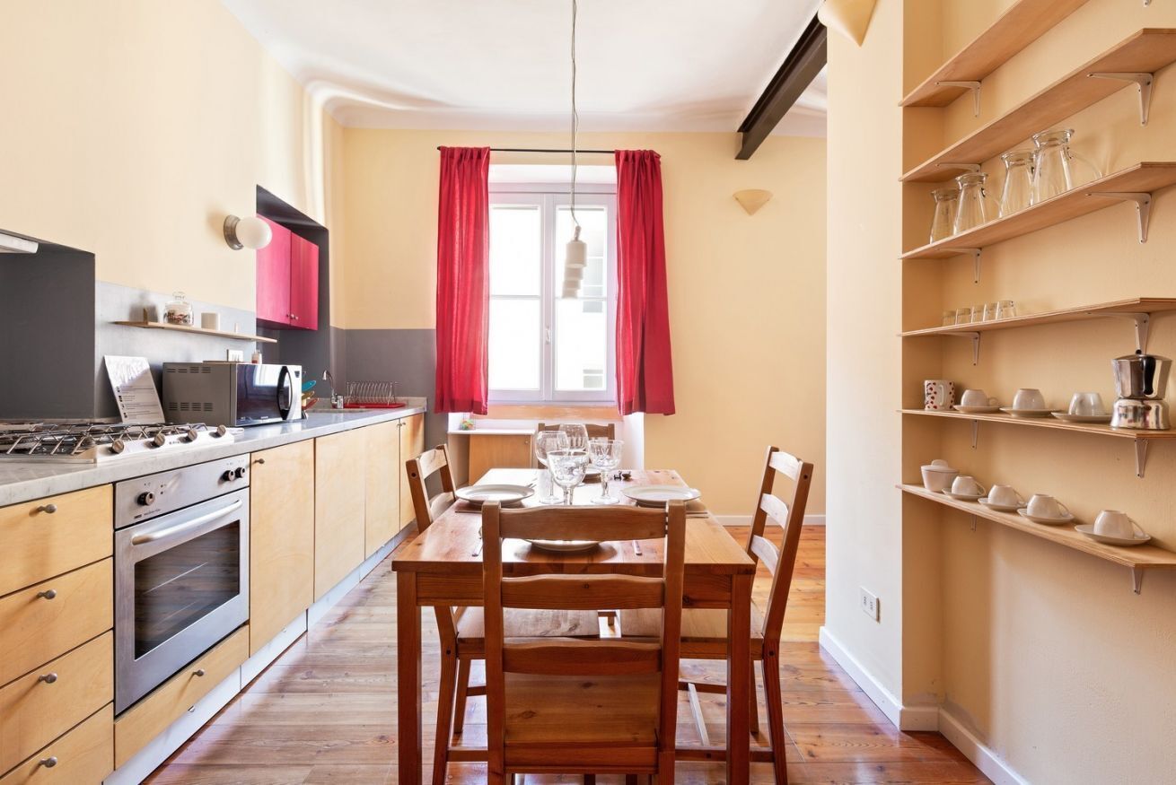 Una cucina con tavolo da pranzo per quattro persone, mobili in legno, forno a incasso e una finestra con tende rosse.