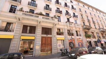 Un edificio a più piani con un ingresso centrale, balconi e veicoli parcheggiati sulla strada.