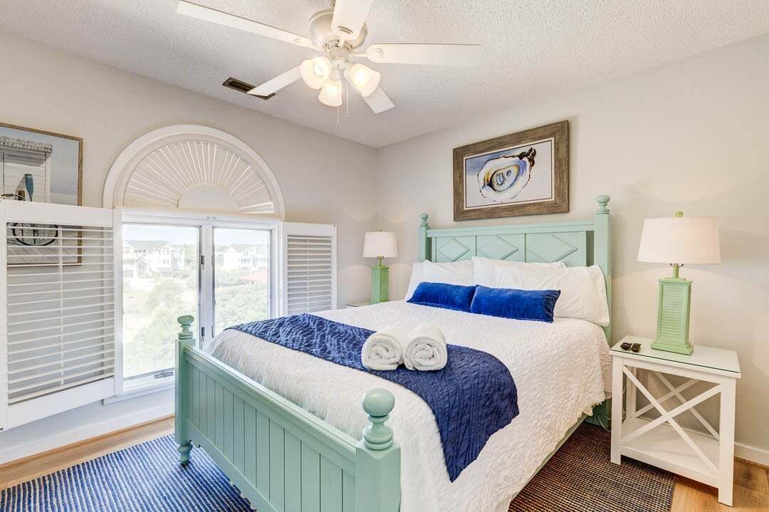 A bedroom with a bed, a ceiling fan, a window with blinds, a framed picture on the wall, and a nightstand with a lamp.