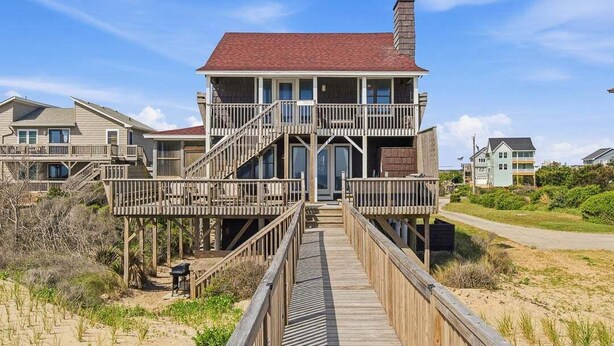 A multi-story house with a balcony, a wooden deck, and a staircase leading to the entrance. - The Porch Life | Oceanfront | Hot Tub | Nags Head (Nags Head)
