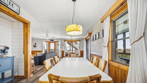 A dining area with a long table, wicker chairs, a hanging light fixture, and a view through a window. - The Porch Life | Oceanfront | Hot Tub | Nags Head (Nags Head)