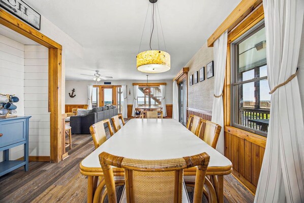 A dining area with a long table, wicker chairs, a hanging light fixture, and a view through a window.