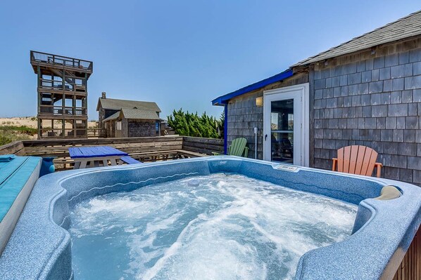 A hot tub in a backyard with a wooden deck, a wooden structure, and a house with a blue roof.