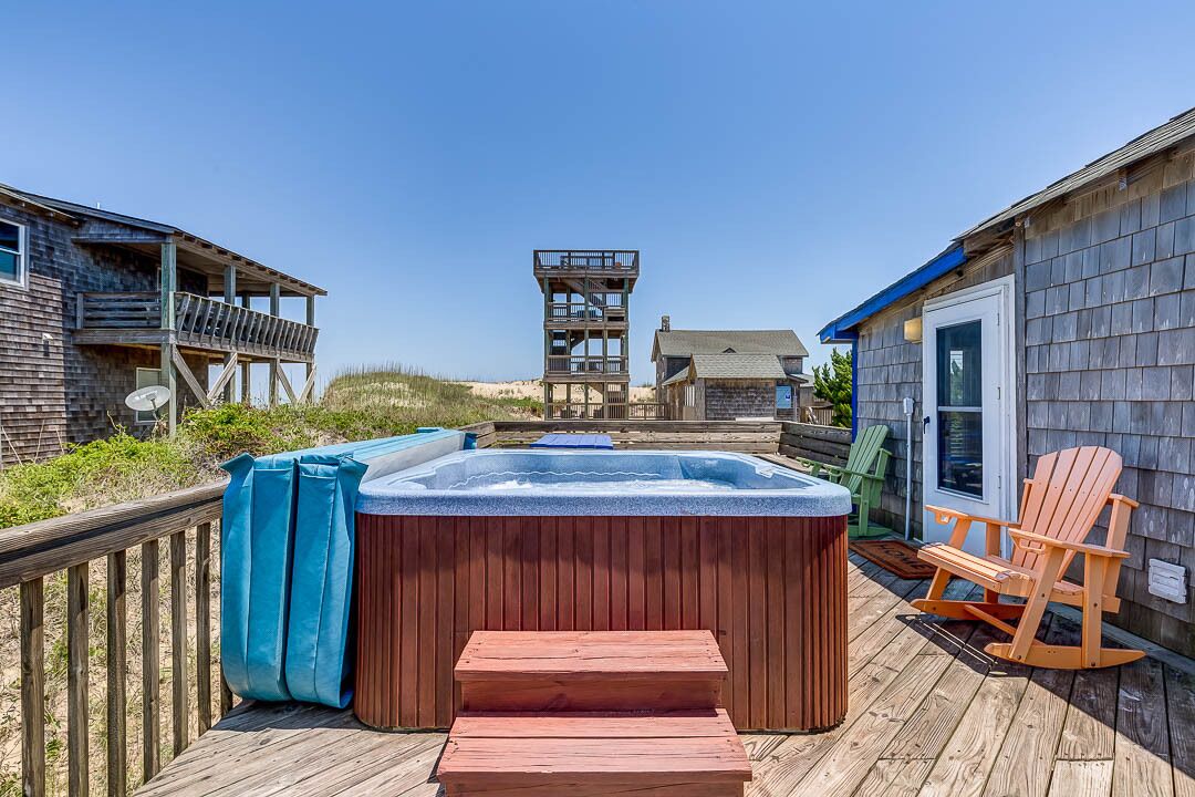 Une terrasse en bois avec un jacuzzi, une chaise en bois et une vue sur une plage et un grand bâtiment en arrière-plan.