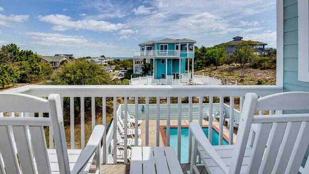 Une maison turquoise à deux étages, dotée d’un balcon donnant sur une piscine, et entourée de verdure et d’autres maisons.