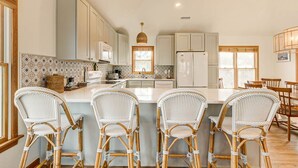 Une cuisine moderne avec des meubles blancs, un îlot central doté de tabourets de bar et un coin repas avec des chaises en bois.