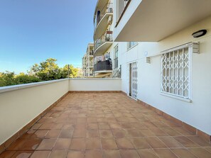 Een balkon met woonfunctie, een betegelde vloer, een witte reling en uitzicht op omliggende gebouwen.