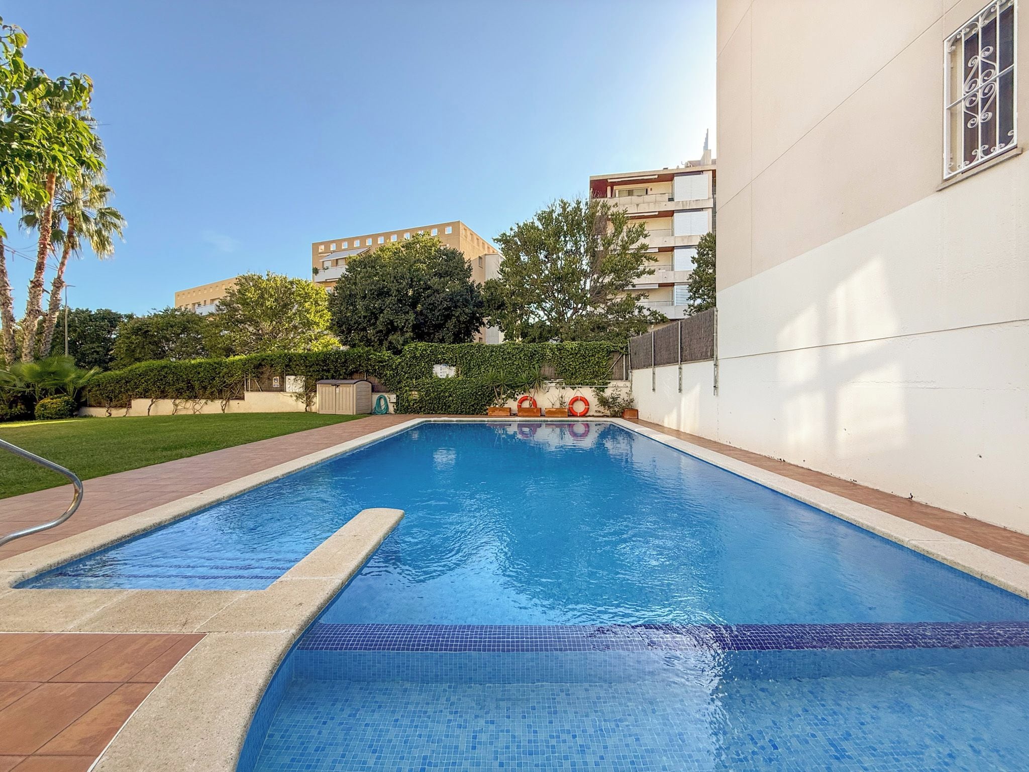 A swimming pool with a tiled edge, surrounded by a paved walkway, and adjacent to a building with a white exterior.