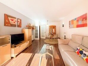 Een moderne woonkamer met een flatscreen televisie, houten kasten, een glazen salontafel, een beige bank en een eethoek met een kast en stoelen.