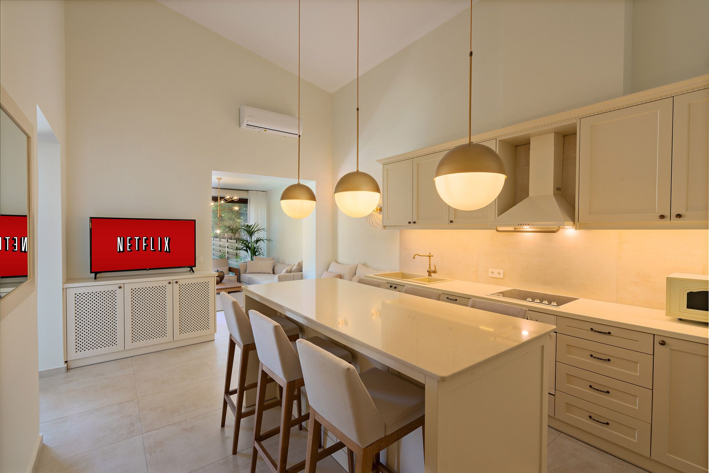 Una cocina moderna con una isla central, taburetes de bar, iluminación colgante y un televisor sobre un mueble.