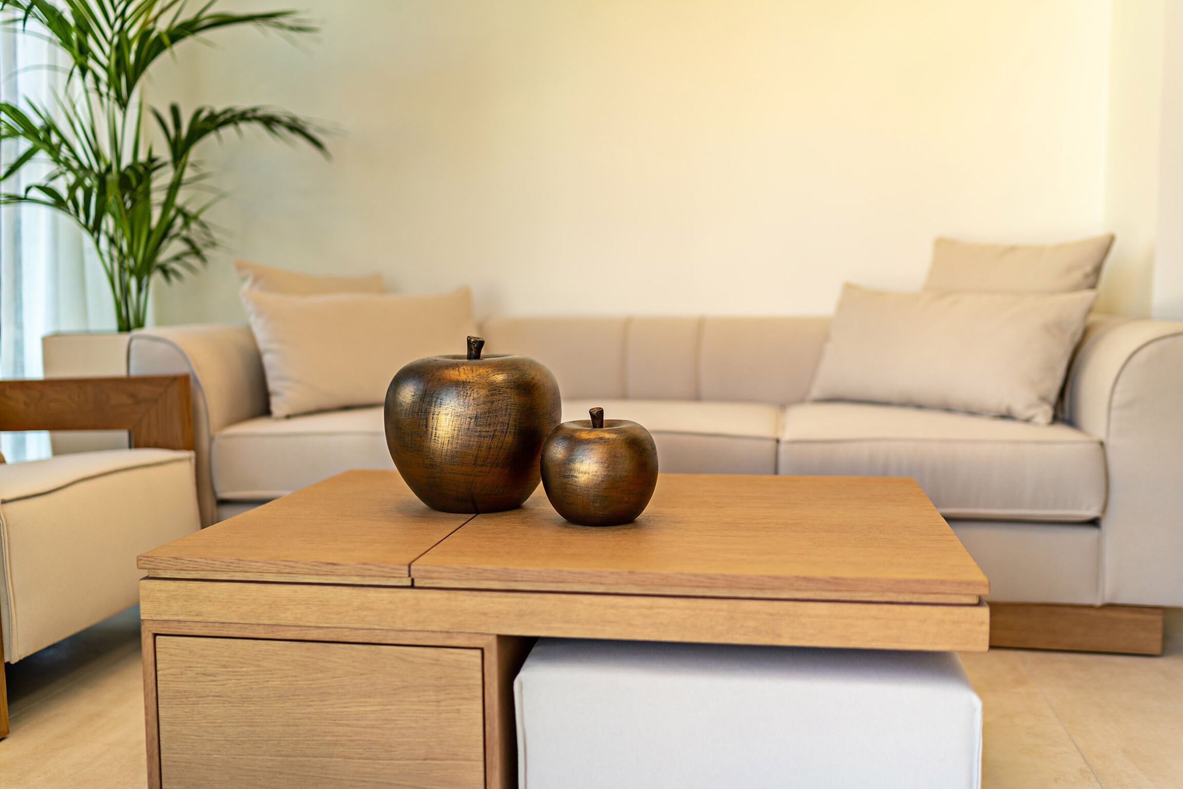 A modern living room with a wooden coffee table, two metallic apples, a beige sofa, and a potted plant.