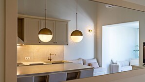 A modern kitchen with a central island, pendant lighting, and a view into a living area with a sofa and a television. - 100m from the beach, kids pool, ping pong, sauna & gym, private sun beds! (Rethymno)