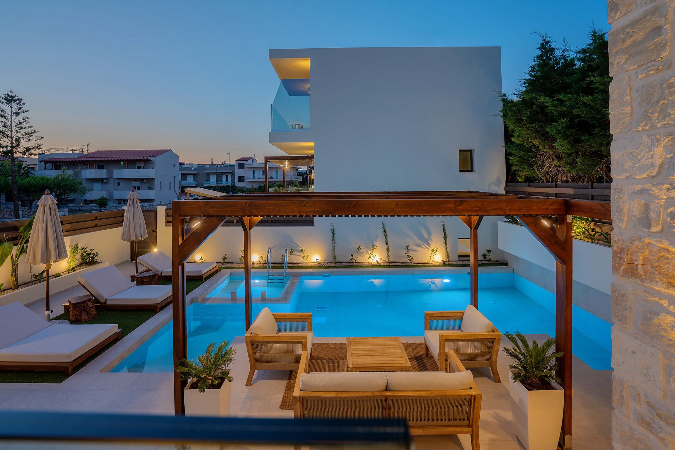Un espace piscine extérieur moderne, doté de chaises longues et de parasols, entouré de murs en pierre et agrémenté d’une pergola en bois.