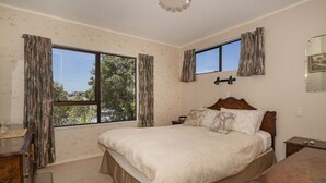 A bedroom with a large bed, two windows with curtains, a bedside table, and a dresser. - Jacaranda Lodge - Whangamata Upstairs Unit (Whangamata)