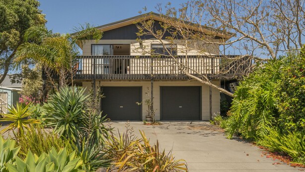 A two-story house with a balcony, garage doors, and a variety of plants in the driveway. - Jacaranda Lodge - Whangamata Upstairs Unit (Whangamata)