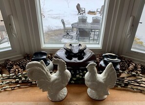 Una ventana con vistas a un paisaje nevado, una mesa con sillas, y gallinas de cerámica y piñas decorativas en el alféizar.