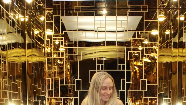 A woman standing in a hallway with mirrored walls and geometric patterns.