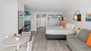 Une chambre d’hôtel moderne équipée d’un lit, d’un canapé, d’une table à manger et offrant une vue sur une plage tropicale par la fenêtre.