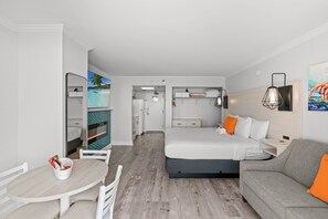 A modern hotel room with a bed, a sofa, a dining table, and a view of a tropical beach through a window.