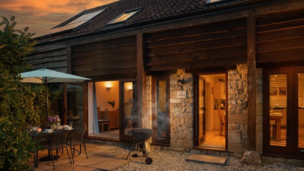 Una casa rústica de madera con fachada de piedra, un patio con mesa y sillas, y una parrilla.