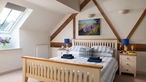 Un dormitorio con techo inclinado, una cama con ropa de cama a rayas, una mesita de noche con una lámpara, una mesita auxiliar con una botella y un vaso, y una ventana con flores.