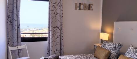A bedroom with a bed, pillows, a bedside table with a lamp, a chair, and a view of the sea through the window.