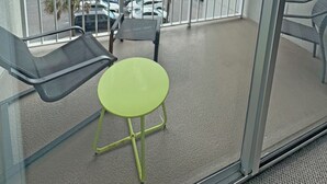 A balcony with a green chair, a black chair, and a table. In the background, there is a view of a parking lot with cars and a building. - Spectacular OCEAN VIEWS - Fabulous Kingston South Hampton 3rd fl unit (Myrtle Beach)